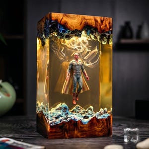 May include: A rectangular resin sculpture featuring a superhero figure suspended within. The sculpture has a wooden base and top with blue and gold accents. The figure is wearing a blue and gold costume with a red face and cape.