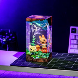 Puede incluir: Una pieza de arte rectangular de resina con personajes de Animal Crossing. Tiene una base de madera y una parte superior con detalles azules y blancos. En el interior, hay figuras de un oso, un conejo y una peque&ntilde;a casa, con follaje verde y una fuente de luz.