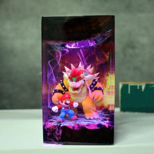 Op de afbeelding: Een rechthoekig kunstwerk van hars met de personages Mario en Bowser uit de Super Mario-videogame serie. Bowser is een groot, geel, schildpadachtig wezen met stekels, en Mario draagt zijn klassieke rode en blauwe outfit. De achtergrond heeft een paarse en oranje gloed.