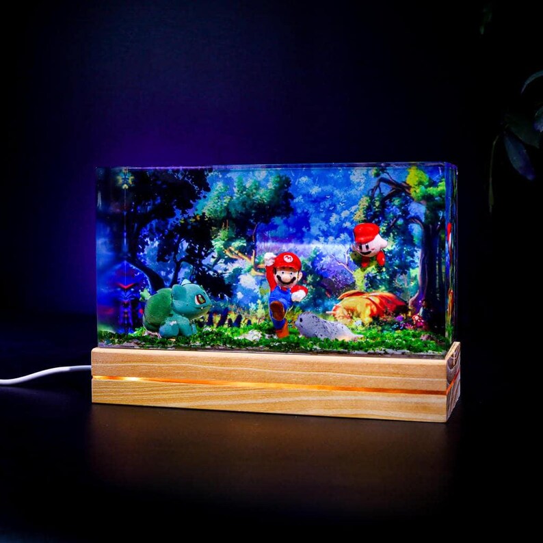 Puede incluir: L&aacute;mpara rectangular de resina con figuras de Mario, Luigi y Bulbasaur en un entorno forestal. La l&aacute;mpara tiene una base de madera con luz c&aacute;lida y un cable de alimentaci&oacute;n. La escena est&aacute; dentro de un bloque de resina transparente.