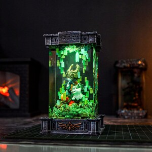 May include: A detailed, illuminated diorama featuring figures from a fantasy game. The scene is encased in a clear, rectangular structure with a castle-like top and base. Green pixelated elements and figures are visible inside, with a green glow emanating from within.