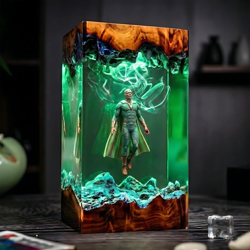 May include: A rectangular resin art piece featuring a superhero figure suspended within. The piece has a wooden base and top, with a vibrant green interior. The figure is in a green and gold costume, with a cape. The resin has a swirling, ethereal effect.