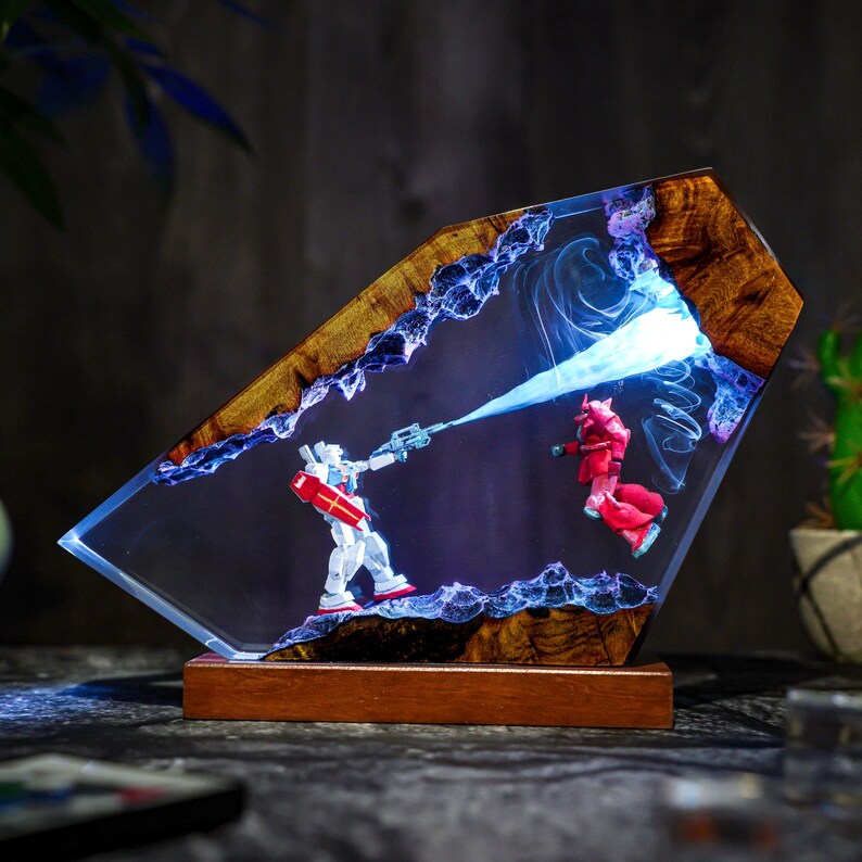 Puede incluir: Una pieza de arte de resina &uacute;nica que presenta dos figuras de robots en una escena de batalla. La obra de arte est&aacute; encapsulada en una resina transparente con un marco y una base de madera. Las figuras son blancas y rojas, con un efecto de luz azul.