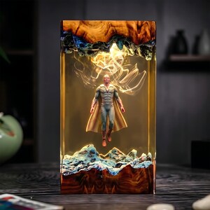 May include: A rectangular resin art piece featuring a superhero figure suspended within. The resin block has a wooden base and top with blue and white accents. The figure is wearing a blue and gold costume with a cape.