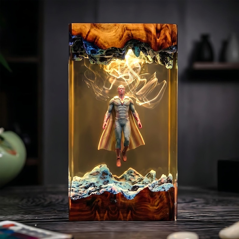 May include: A rectangular resin art piece featuring a superhero figure suspended within. The resin block has a wooden base and top with blue and white accents. The figure is wearing a blue and gold costume with a cape.