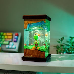 May include: A decorative lamp featuring a green Yoshi figurine inside a clear resin cube. The lamp has a wooden frame and base, with a landscape design. The interior includes a green base, red mushrooms, and a blue mountain scene.