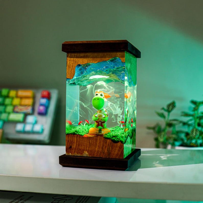 May include: A decorative lamp featuring a green Yoshi figurine inside a clear resin cube. The lamp has a wooden frame and base, with a landscape design. The interior includes a green base, red mushrooms, and a blue mountain scene.