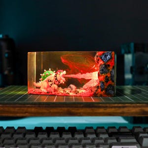 May include: A resin diorama featuring a Bowser figure breathing fire. The scene is encased in a clear rectangular block, with red and orange lighting effects. The figure has green spikes and a black studded collar.