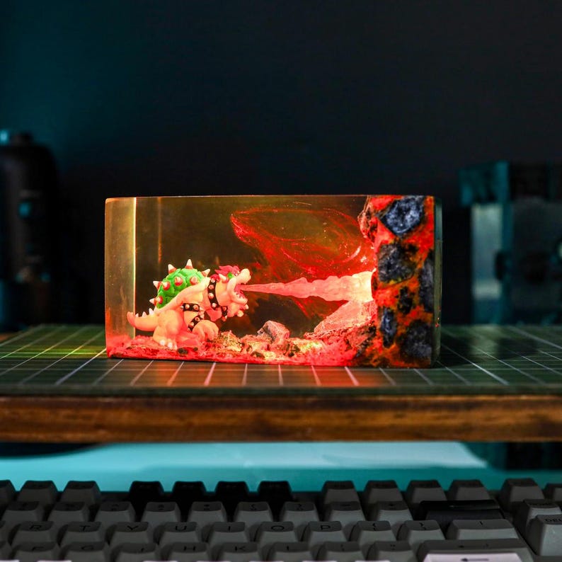 May include: A resin diorama featuring a Bowser figure breathing fire. The scene is encased in a clear rectangular block, with red and orange lighting effects. The figure has green spikes and a black studded collar.