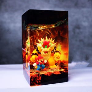 Op de afbeelding: Een rechthoekige decoratieve doos met een levendige afbeelding van de videogamepersonages Mario en Bowser tegen een vurige achtergrond. De doos is voornamelijk zwart met de kleurrijke personages en achtergrondkunst die rond de zijkanten loopt.