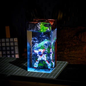 Pu&ograve; includere: Un'opera d'arte in resina rettangolare con una figura di robot bianca, rossa e blu racchiusa in un blocco trasparente tinto di blu. La parte superiore presenta elementi verdi e marroni, con uno sfondo scuro.