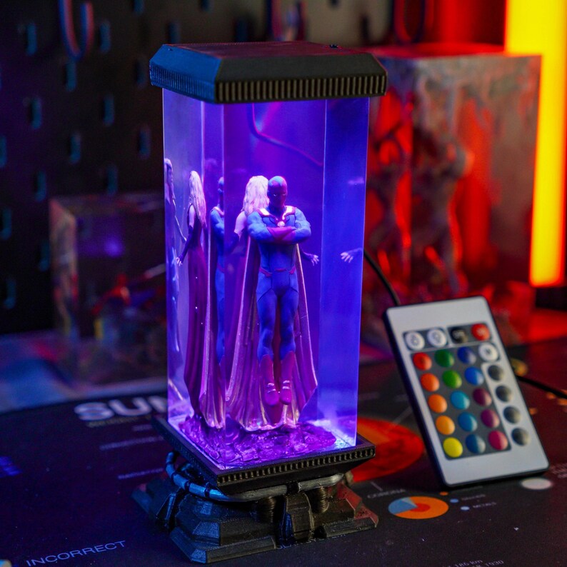 Puede incluir: Una vitrina rectangular iluminada con figuras de superh&eacute;roes. La vitrina tiene una base y una parte superior negras, con una secci&oacute;n central transparente. Un mando a distancia con opciones de color est&aacute; cerca.