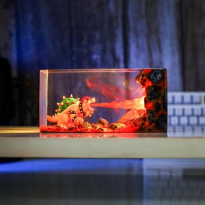 May include: A resin sculpture of Bowser breathing fire, set within a clear rectangular block. The design includes volcanic rock and lava. The Bowser figure is orange, green, and black, creating a dynamic scene.