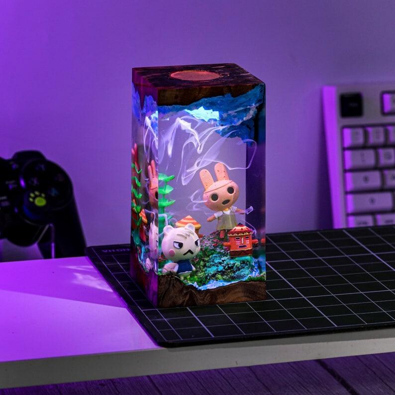 May include: A rectangular resin lamp featuring characters from Animal Crossing. The lamp has a wooden top and base, with a colorful scene inside. The characters include a rabbit, a cat, and a small red structure. The lamp is lit with a soft, diffused light.