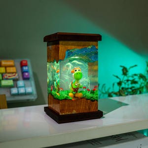 May include: A wooden and resin lamp featuring a green cartoon character in a landscape with red mushrooms. The lamp has a rectangular shape with a dark brown wooden frame and a clear resin center. The scene is illuminated with a soft, warm light.