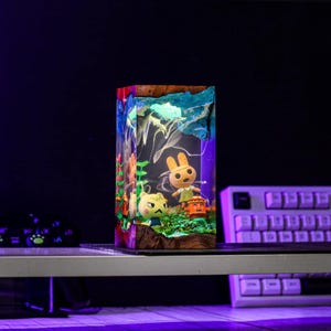 Puede incluir: L&aacute;mpara rectangular de resina con personajes de Animal Crossing. La l&aacute;mpara tiene una base de madera y una parte superior azul y blanca, con figuras coloridas en el interior. La l&aacute;mpara se ilumina con un brillo p&uacute;rpura y azul, creando un efecto caprichoso.