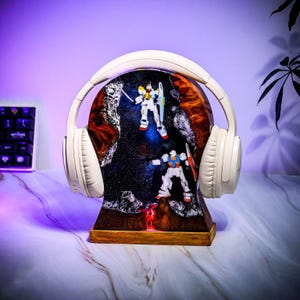 May include: A white headset rests on a wooden headphone stand with a resin design featuring two robot figures in a starry space. The stand has a wooden base and a warm, glowing light.