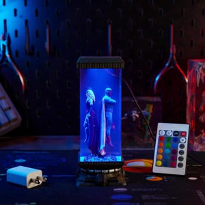 Puede incluir: Una l&aacute;mpara cil&iacute;ndrica iluminada en azul, con figuras de una mujer con un vestido oscuro y un hombre con un disfraz de superh&eacute;roe. La l&aacute;mpara tiene una base y una parte superior negras, con un mando a distancia. Tambi&eacute;n se ve un adaptador de corriente blanco.