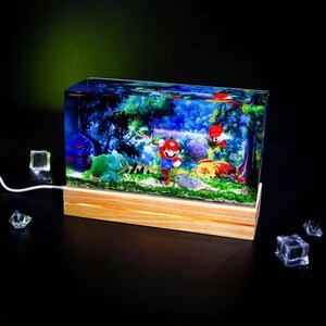 Puede incluir: Una luz nocturna rectangular de resina con un diorama en miniatura de una escena de videojuego. La escena incluye figuras de un personaje con sombrero rojo, un dinosaurio verde y otros personajes. La luz se asienta sobre una base de madera y tiene un cable de alimentaci&oacute;n blanco.