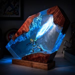 May include: A unique resin lamp featuring a detailed figurine of a character from a video game. The lamp has a wooden base and is encased in a clear resin with blue accents, creating a glowing effect. The top is made of wood.