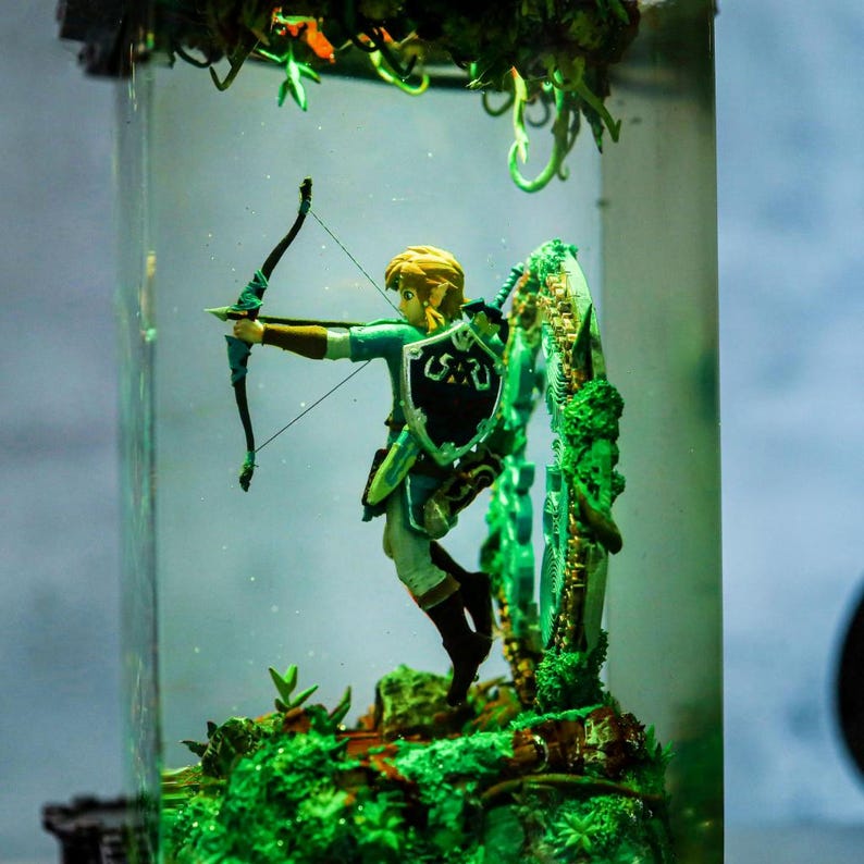 以下が含まれることがあります： ゼルダの伝説のキャラクターのフィギュアで、弓矢を構えています。緑と白の衣装を着ており、紋章の盾を持っています。透明な円筒形の容器の中にあり、緑の葉で囲まれています。