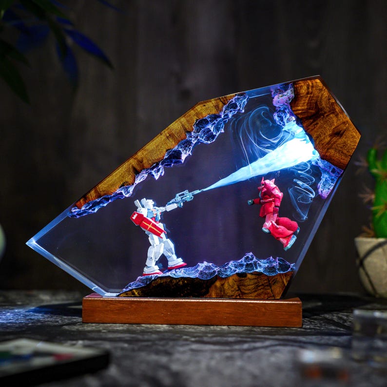 Puede incluir: Una pieza de arte de resina &uacute;nica que presenta dos figuras de robots en una escena de batalla. La obra de arte est&aacute; encapsulada en una resina transparente con un marco y una base de madera. Las figuras son blancas y rojas, con un efecto de luz azul.
