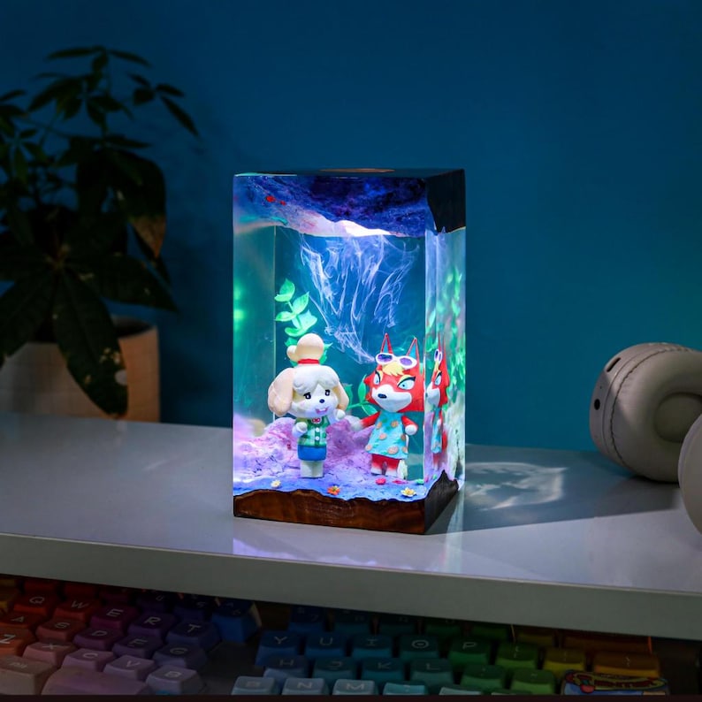 May include: A rectangular resin lamp featuring two cartoon characters inside a clear block. The lamp has a wooden base and a dark top, with blue and green lighting effects. The characters are a dog and a fox, both wearing colorful outfits.