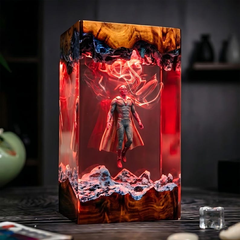 May include: A rectangular resin art piece featuring a superhero figure suspended within. The resin is a mix of amber, blue, and white, with a red glow emanating from within. The superhero is wearing a gray and red costume.
