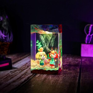 May include: A rectangular resin art piece featuring two cartoon characters, Isabelle and Redd, from Animal Crossing. The figures are set in a clear resin block with a dark wooden base, with a colorful, glowing effect.