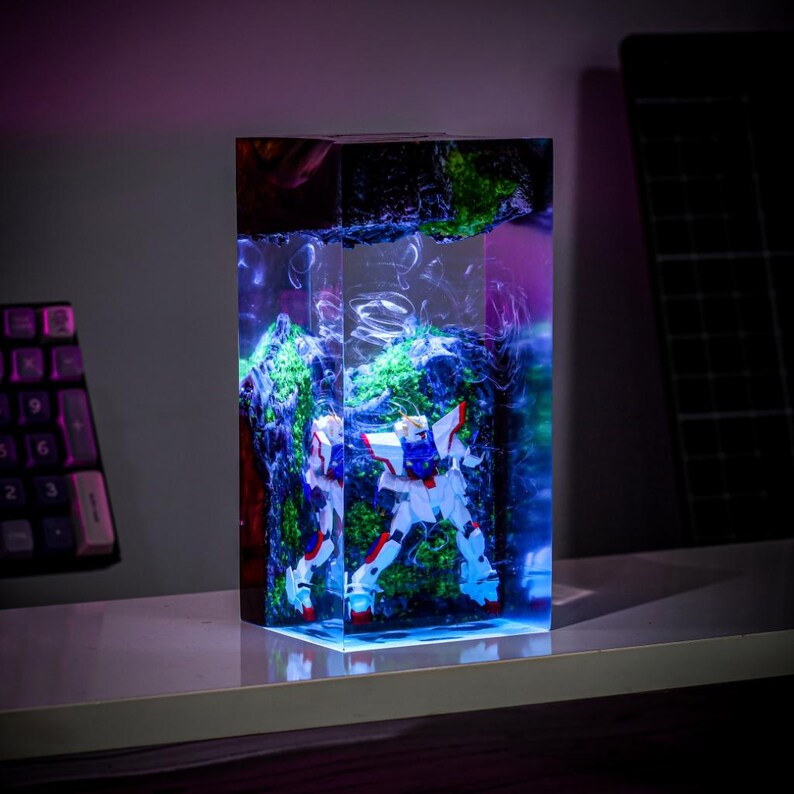 Pode incluir: Escultura retangular em resina com uma figura de rob&ocirc; branco, azul e vermelho contra um fundo de elementos texturizados verdes e azuis. A escultura &eacute; iluminada, criando uma exibi&ccedil;&atilde;o vibrante. A pe&ccedil;a &eacute; exibida em uma superf&iacute;cie branca.