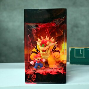 Op de afbeelding: Een rechthoekige displaycase met de personages Mario en Bowser uit de Super Mario-videogameserie. Bowser is een groot, geel schildpadachtig wezen met stekels, en Mario is een klein figuur met een rode hoed en blauwe overall. De achtergrond is vuurrood.