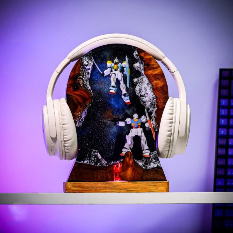 May include: White wireless headphones framing a resin art piece featuring two robot figures in a space-themed setting. The art piece has a wooden base and a galaxy-like background with red and blue hues.