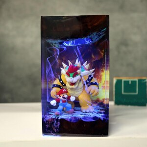 Op de afbeelding: Een rechthoekig harskunstwerk met de personages Mario en Bowser uit de Super Mario-videogameserie. Bowser is een groot, geel schildpadachtig wezen met stekels, en Mario is een klein figuur in rood en blauw. De achtergrond heeft een vurig, blauw verlicht ontwerp.