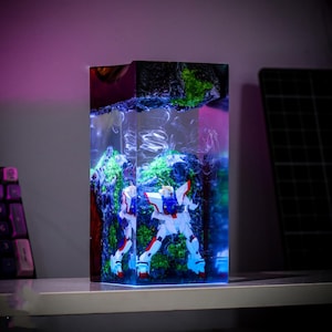 Pu&ograve; includere: Un'opera d'arte rettangolare in resina con una figura di robot bianco, rosso e blu all'interno. Il blocco di resina ha un design a forma di montagna con muschio verde e una base luminosa blu. Il pezzo &egrave; su una mensola bianca.