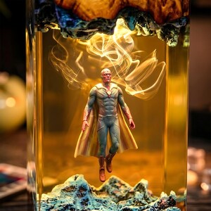 May include: A figurine of a superhero with a red face, green and blue suit, and a yellow cape, suspended in a clear, rectangular container. The container has a yellow-orange liquid inside, with a decorative base and top. The background is blurred.