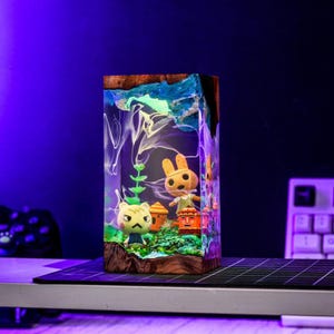 May include: A rectangular resin art piece featuring miniature figurines of characters from the Animal Crossing video game. The artwork has a wooden base and a blue and white top, with the characters and scenery inside. The piece is illuminated with a soft, glowing light.