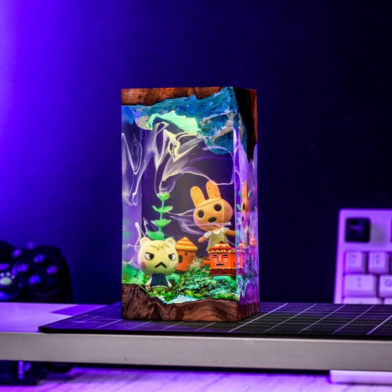 May include: A rectangular resin art piece featuring miniature figurines of characters from the Animal Crossing video game. The artwork has a wooden base and a blue and white top, with the characters and scenery inside. The piece is illuminated with a soft, glowing light.