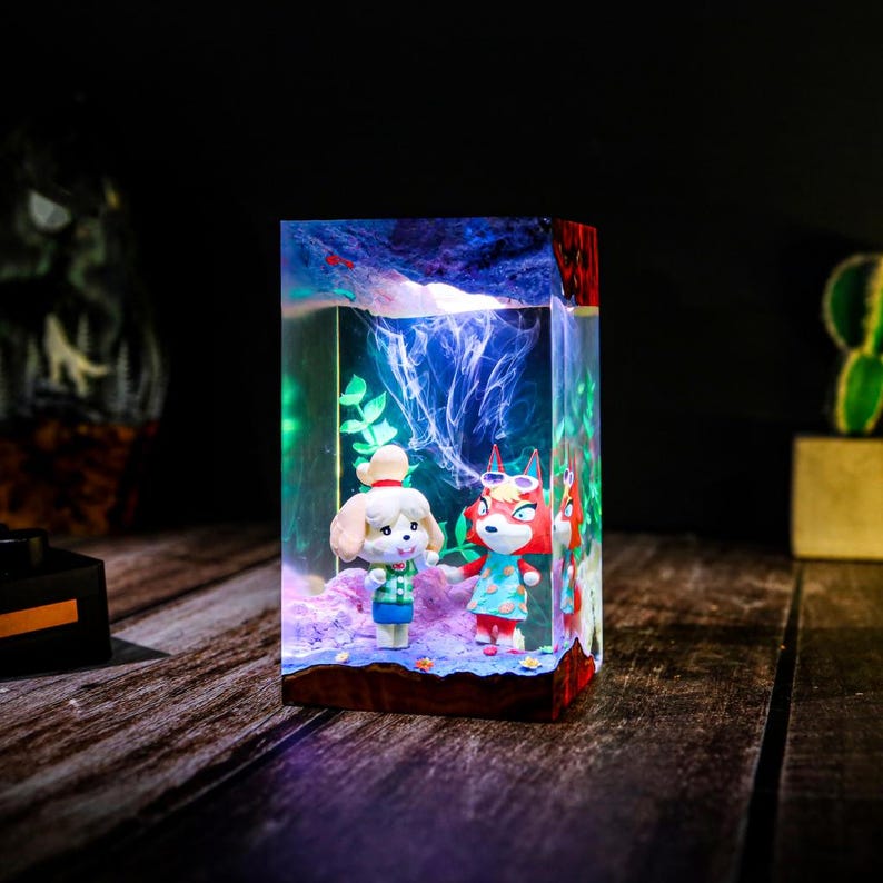 May include: A rectangular resin art piece featuring two cartoon characters, one a dog and the other a fox, standing in a miniature landscape. The piece has a wooden base and a light inside, creating a glowing effect. The background has a blue and white design.