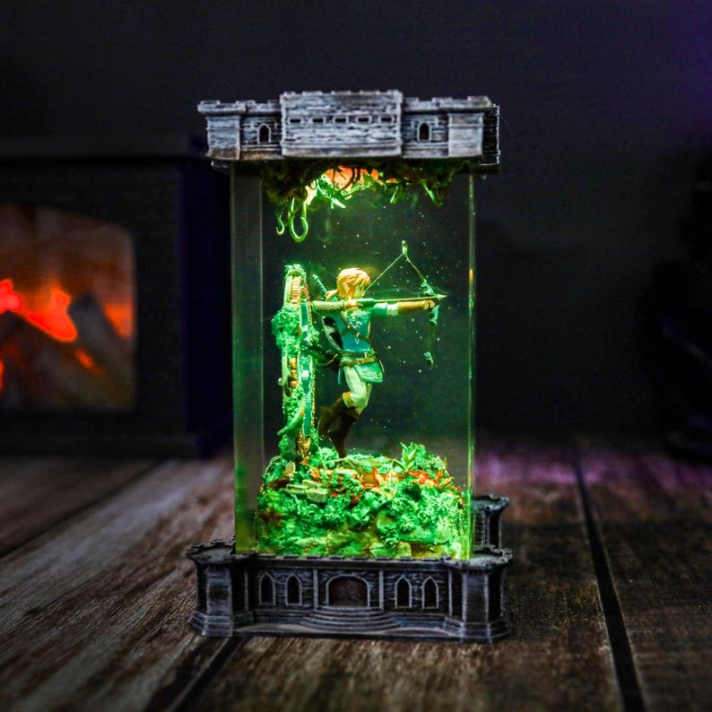 以下が含まれることがあります： 透明な長方形のディスプレイケースの中に、弓矢を持ったキャラクターの精巧なフィギュア。フィギュアは緑と茶色の衣装を着ており、緑色の台座の上に立っています。ケースは灰色の城のような構造で囲まれ、緑色の光がシーンを照らしています。