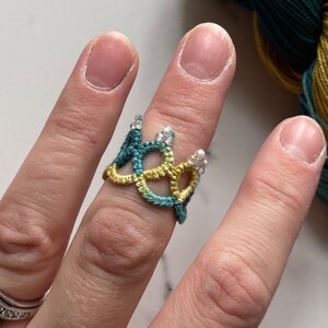 May include: A green, yellow and white crocheted ring with small white beads. The ring is worn on a finger.