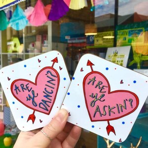 May include: Two white square coasters with pink hearts and red arrows. The text on the coasters reads "Are ye dancin?" and "Are ye askin'?"