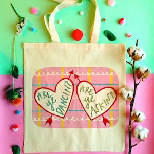 May include: A white tote bag with pink and yellow hearts that say "Are ye dancin?" and "Are ye askin?" on a pink and yellow plaid background.