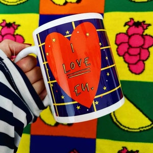 May include: White ceramic mug with a blue and white checked pattern and a red heart with the text "I LOVE EU" in black. The mug is held by a hand wearing a striped shirt.