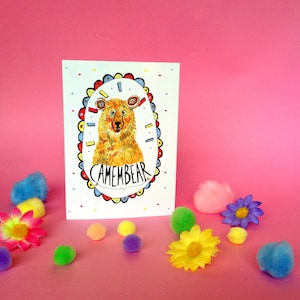 May include: A white card with a colorful illustration of a bear with the text "Camembear" inside a circle. The card is surrounded by colorful pom-poms on a pink background.