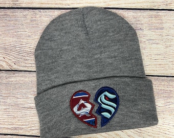 May include: A gray beanie with a broken heart design. One half of the heart features the Colorado Avalanche logo, and the other half features the Seattle Kraken logo. The hat is made of a soft, knitted material.