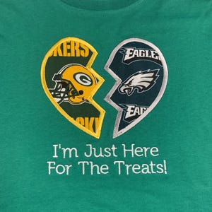 May include: Green t-shirt with a broken heart design featuring the Green Bay Packers and Philadelphia Eagles logos. The text "I'm Just Here For The Treats!" is embroidered below the design.
