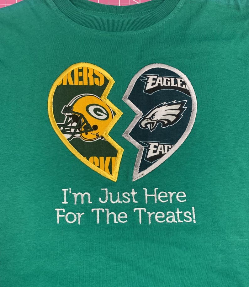 May include: Green t-shirt with a broken heart design featuring the Green Bay Packers and Philadelphia Eagles logos. The text "I'm Just Here For The Treats!" is embroidered below the design.