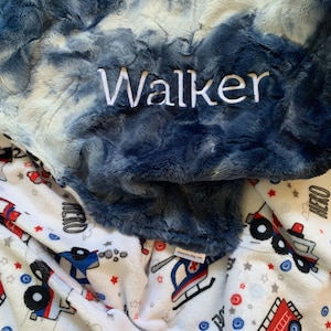 May include: A plush, navy blue and white tie-dye baby blanket with the name "Walker" embroidered in white. The blanket is layered over a white blanket printed with red, blue, and gray fire trucks, helicopters, and stars.