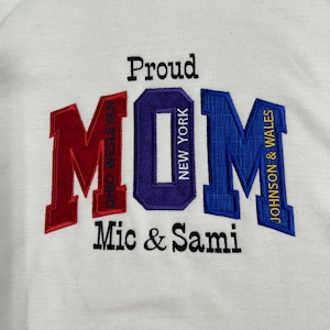 May include: A white hooded sweatshirt featuring embroidered text. The word Proud is in black, and MOM is in large red, purple, and blue letters. Inside the letters are the words Ohio Wesleyan, New York, and Johnson & Wales. Mic & Sami is in black.