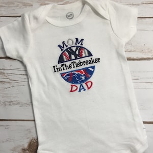 May include: White baby onesie with a baseball-themed design. The design includes the words "MOM", "I'm The Tiebreaker", and "DAD" in blue and red embroidery. The onesie has short sleeves and snap closures.